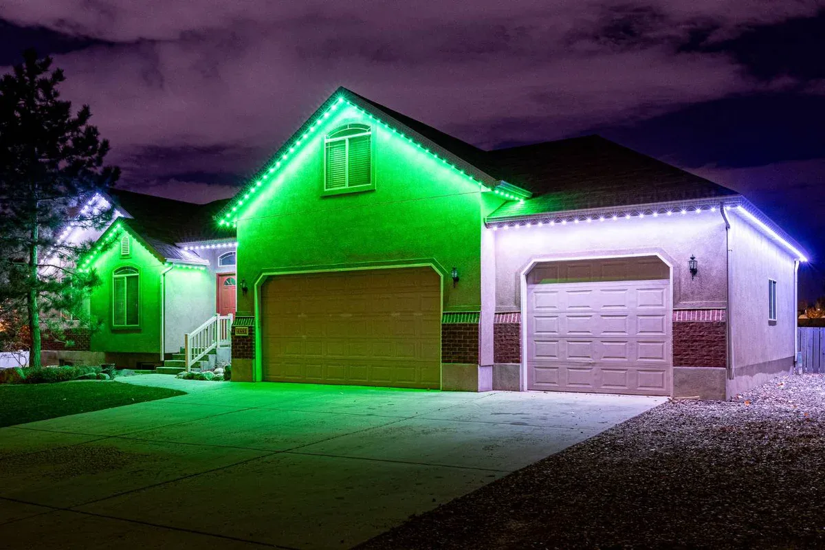 Attached image file A suburban house at night features green and white LED lights along the roofline, illuminating the garage doors and front entrance. The driveway and sky are visible, with some clouds overhead.