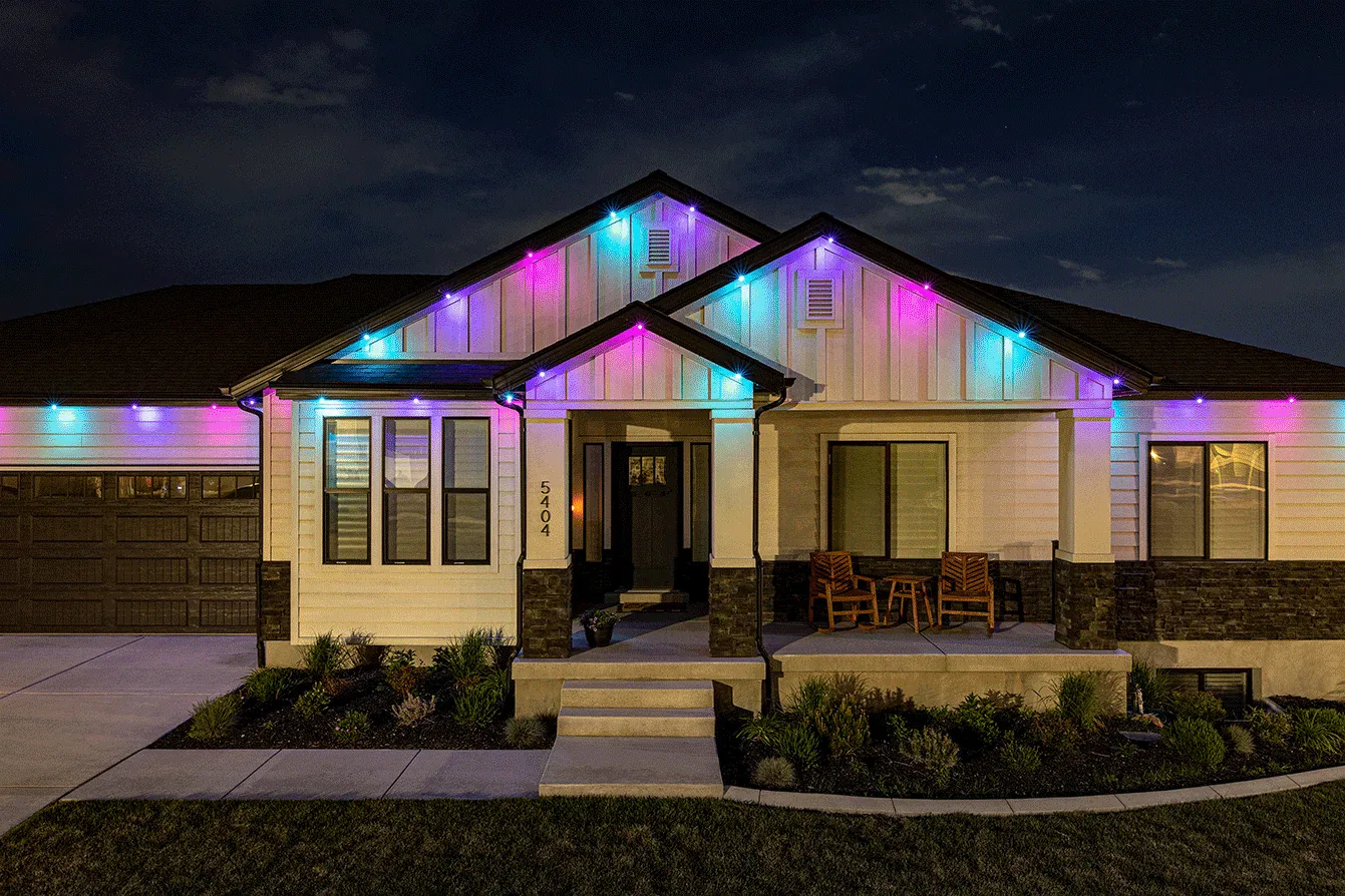 Attached image file A single-story house at night displays colorful lights in blue, pink, and purple along the roofline. The porch has two wooden chairs and the yard is neatly landscaped.