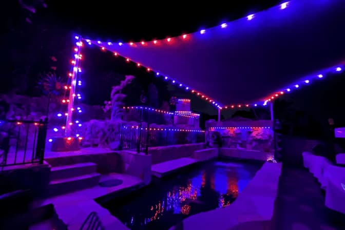 Attached image file A backyard pool at night illuminated by red and blue string lights along a canopy and fence, with reflections of the lights shimmering on the water. The surrounding area appears tranquil and festive.