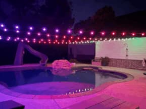 A backyard pool at night is lit with colorful string lights in purple, red, and white. The pool has a slide, rock feature, outdoor shower, and seating, creating a festive and inviting atmosphere.