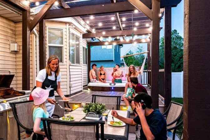 Attached image file A group of people enjoy a backyard gathering; some are eating at a table under string lights while others relax in a hot tub on a raised deck, creating a lively, outdoor evening scene.