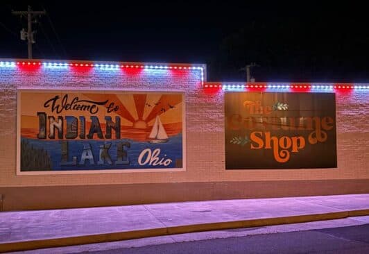 Attached image file A brick building at night with two large murals: one reads Welcome to Indian Lake Ohio with a lake scene, the other says The Costume Shop in orange letters. Red, white, and blue lights outline the roof.