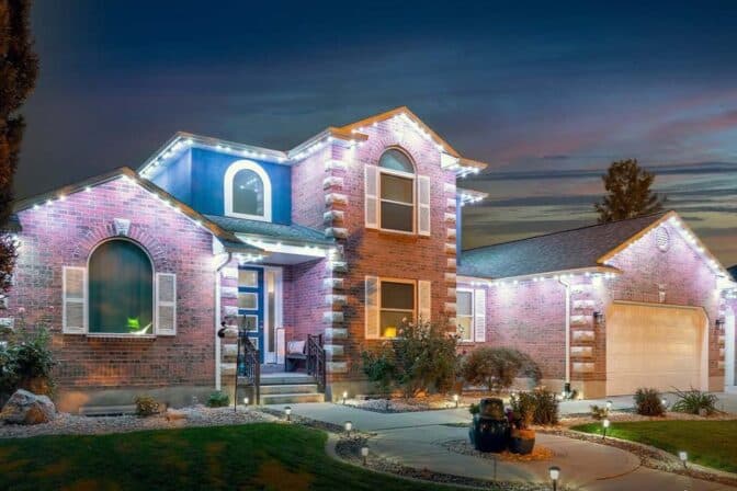 A two-story brick house is illuminated by bright white lights along the roofline at dusk, with shrubs and landscaping in the front yard and a double garage on the right side.