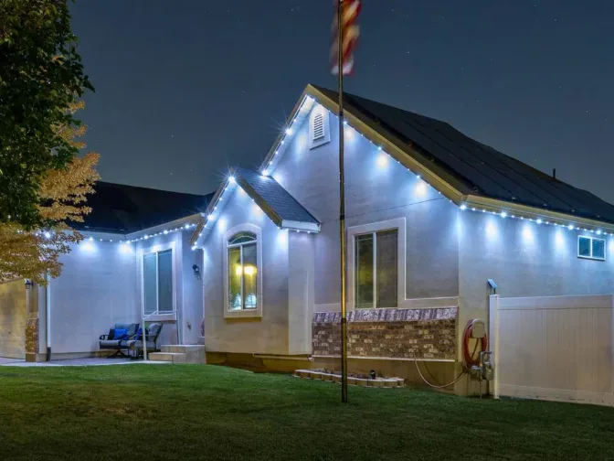 A single-story house at night with white exterior walls, outdoor string lights along the roofline, a flagpole in front, a manicured lawn, and a tree partially visible on the left.