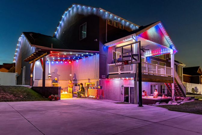 Attached image file A house at dusk decorated with colorful string lights, featuring a balcony, basketball hoop, and people gathered on the patio and under the covered area in the backyard.