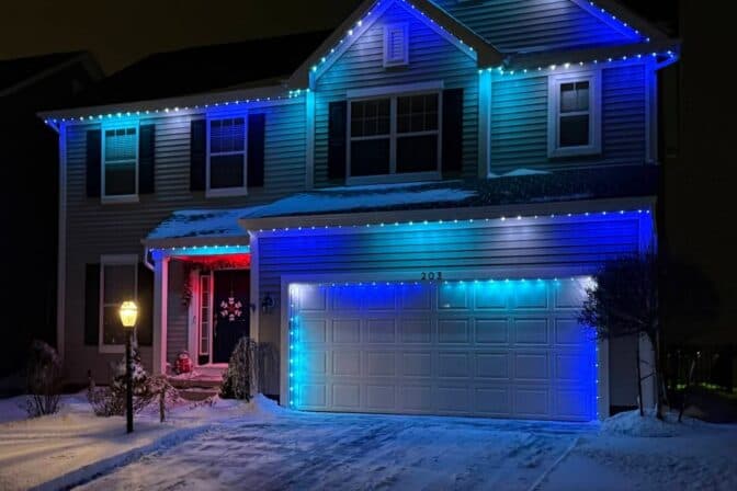 Attached image file A two-story house is decorated with blue and white lights along the roofline at night, with snow covering the yard, trees, and driveway. A lit lamppost stands near the front walkway.