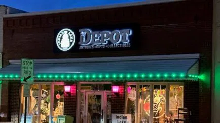 A brick storefront at night with a lit Depot Antiques • Gifts • Collectibles sign, festive window displays, green lights above the awning, and a sidewalk sign that reads Indian Lake Outfitters. A Stop Here on Red sign is nearby.