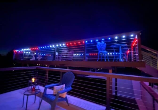 Attached image file A deck at night is decorated with red, white, and blue string lights. There are metal tables and chairs on the upper level and a blue Adirondack chair with a side table and candle on the lower level.
