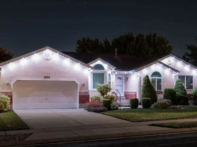 A single-story house at night with white string lights outlining the roof, a two-car garage, neatly trimmed bushes, and a well-kept front yard.