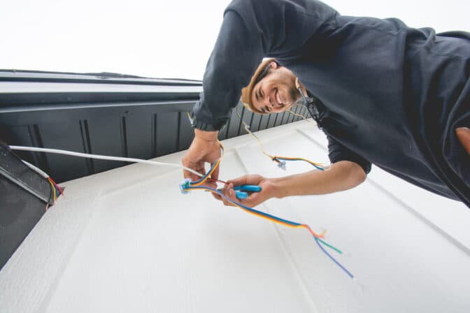 Attached image file A person smiles while working on electrical wiring outside a building, using tools to connect colorful wires near a white wall and dark roof edge.