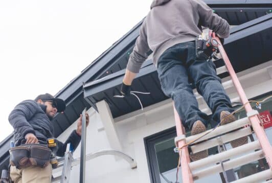 Attached image file Two workers on ladders install or repair wiring under the roof eaves of a house. One holds tools on his belt, while the other manages wires near the roof edge. Both wear casual work clothes and safety gear.