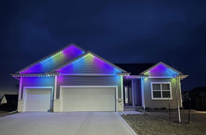 Attached image file Single-story house at night with colorful LED lights outlining the roof, displaying a gradient of blue, purple, and yellow hues; the house has three garage doors and a large driveway.
