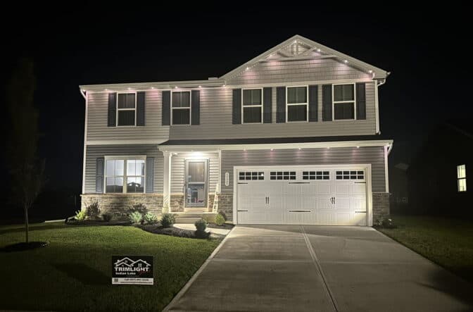 Attached image file A two-story house at night with lights illuminating the exterior. A Trimlight sign is displayed on the front lawn, and the house has a double garage, neatly trimmed grass, and small shrubs by the entrance.