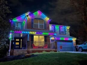 A two-story house decorated with bright, colorful LED lights along the rooflines at night. The lights are in shades of blue, green, pink, and purple, creating a festive look. The yard and driveway are visible in the foreground.