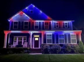 A two-story house at night is decorated with red, white, and blue LED lights outlining the roof and porch, illuminating the front yard and shrubbery.