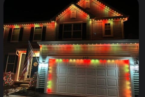 A suburban house is decorated with red, green, and yellow Christmas lights along the roofline and garage. An American flag hangs by the front door, and a small lit tree is in the yard at night.