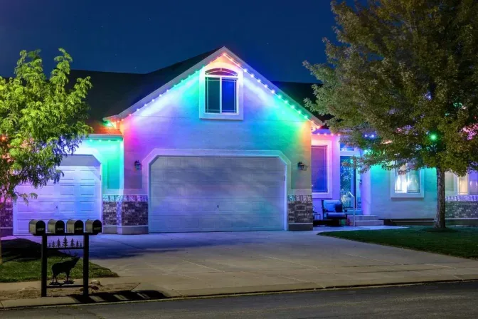 Attached image file Single-story house at night with colorful LED lights outlining the roof and garage. Trees frame the scene, and four mailboxes are visible at the curb. The driveway and front entrance are well lit.