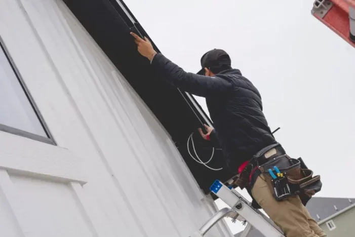 Attached image file A person standing on a ladder installs or repairs electrical wiring under the eaves of a white house, wearing work gear and a tool belt on their waist.