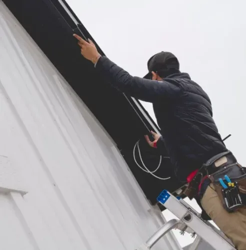 Attached image file A person standing on a ladder installs or repairs electrical wiring under the eaves of a white house, wearing work gear and a tool belt on their waist.