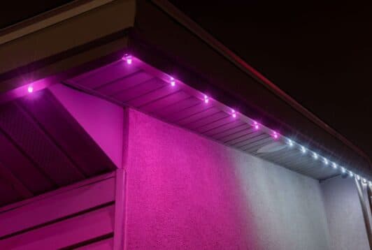 Attached image file The corner of a house roof at night is lit with purple-pink LED lights along the eaves, casting a colorful glow on the wall, with a few white lights visible farther down the edge.