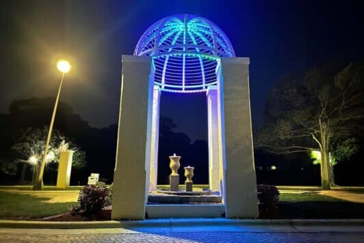 Attached image file A pavilion structure with a domed metal roof glows with blue and white lights at night. Three abstract sculptures stand inside, surrounded by trees and a lit streetlamp in a park-like setting.