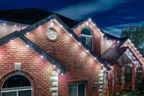 A brick house with arched windows is decorated with white string lights along the roofline, creating a festive look at night under a dark blue sky.