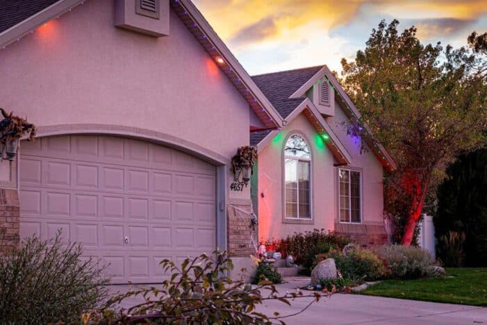 Attached image file A suburban house at sunset with a two-car garage. The house is decorated with colorful holiday lights along the roof and front window. Trees and bushes surround the house, and the driveway is empty.