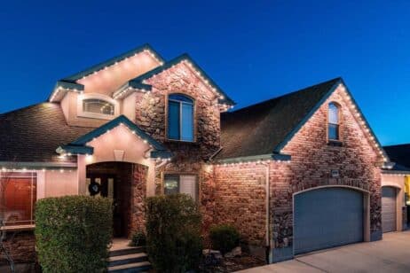 A two-story brick and stone house with a two-car garage, lit by white string lights along the roofline, set against a clear evening sky. Bushes frame the front steps leading to the entrance.