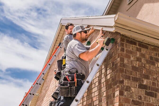 Attached image file Two workers on ladders install or repair a gutter on the roof edge of a brick house. They wear grey shirts and tool belts, working under a partly cloudy sky.