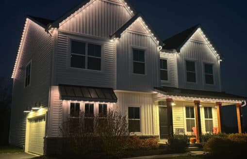 Attached image file A two-story white house at night is illuminated by white string lights along the roofline. The porch has wooden columns, two orange chairs, a pumpkin, and bushes in front. The garage is to the left.