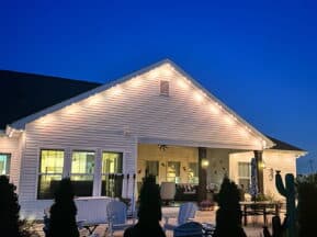 A cozy backyard patio at dusk features white outdoor chairs, a table, potted plants, and string lights illuminating the roofline of a white house against a deep blue evening sky.