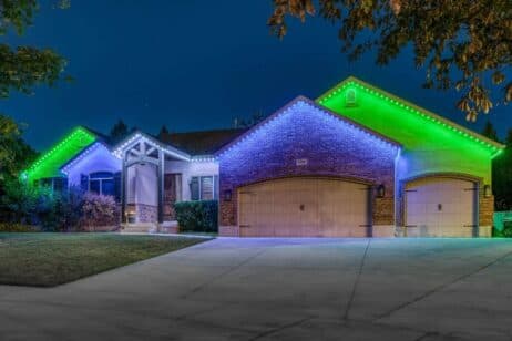A single-story brick house at night with green and white LED lights outlining the roof, three garage doors, and a well-kept front yard with trees and bushes visible.