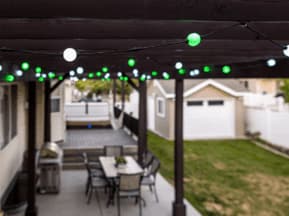 String lights with green and white bulbs hang from a pergola over a patio with a dining table and chairs. In the background, there is a lawn, a barbecue grill, and a detached garage.
