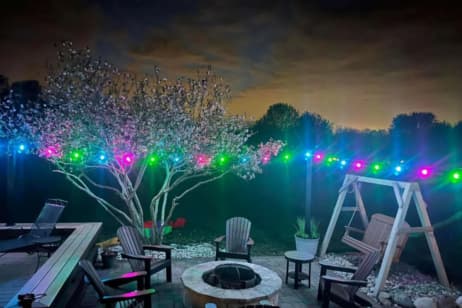 A backyard patio at night features a fire pit surrounded by chairs, a wooden swing, blooming trees, and multicolored string lights, with a cloudy sky illuminated by city lights in the background.