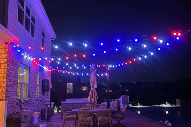 Attached image file A backyard patio at night is decorated with strings of red, white, and blue lights. Patio furniture, including chairs and a closed umbrella, sits near a pool. The house and pool are softly illuminated.