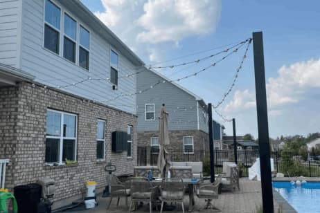 A backyard patio with string lights, outdoor furniture, a large umbrella, and a swimming pool is shown next to a brick house under a partly cloudy sky.