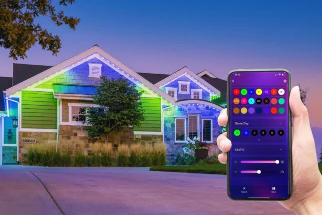 A hand holds a smartphone displaying a color-changing light control app, aimed at a house decorated with vibrant, multicolored LED lights along the roof at dusk.