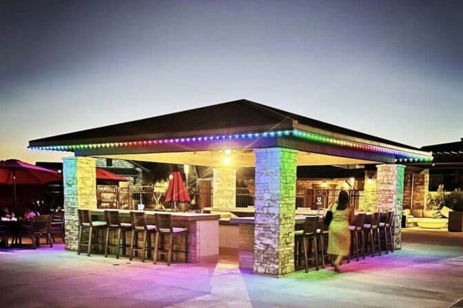 Attached image file Outdoor bar with stone pillars and a dark roof, outlined by colorful string lights at dusk. Barstools surround the bar, and a person is walking nearby. Patio umbrellas and seating are visible in the background.