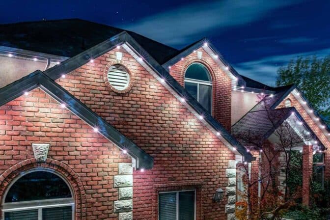 A brick house with gabled roofs is decorated with white outdoor string lights along the roof edges, glowing brightly at night under a clear, starry sky.