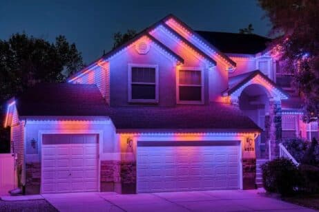 A two-story house at night is decorated with bright purple, pink, and blue LED lights outlining the roof, garage, and entryway, creating a colorful and festive appearance.