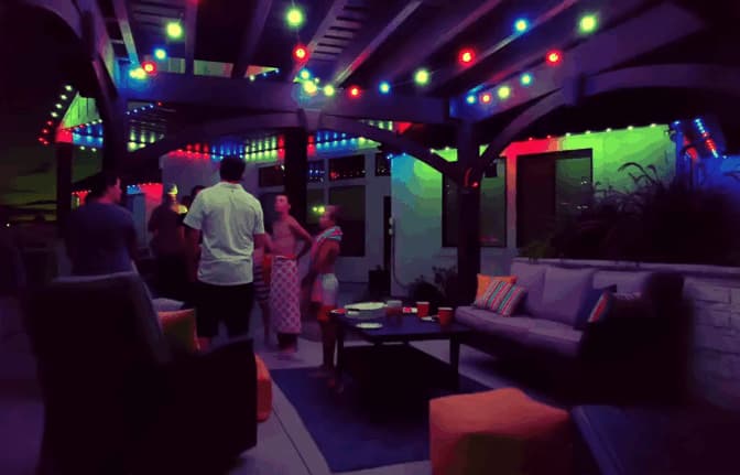 Attached image file A group of people chat and relax under a pergola decorated with colorful string lights at night. Outdoor couches and a table with cups create a cozy, festive atmosphere.