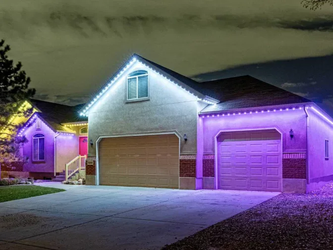 A single-story house with a three-car garage is decorated with white and purple LED lights outlining the roof and garage doors at night. The yard features a tree and a concrete driveway.